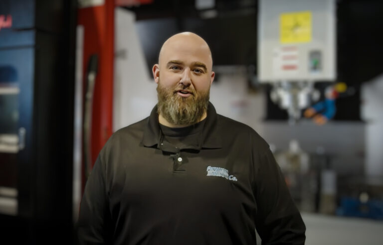 Bearded Ferguson Industrial Co. employee in a black branded polo standing in the machine shop